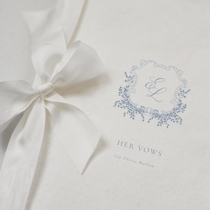 May include: A white wedding vow book tied with a white satin ribbon. The cover features a blue floral design with the text "HER VOWS" and the name "Lia Olivia Marlow".