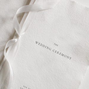 May include: A white wedding program with the text "THE WEDDING CEREMONY" and the names "SAMANTHA & JEREMY". The program is tied with a white ribbon. The paper has a textured finish.