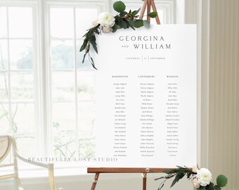 May include: A white wedding seating chart on an easel with a floral arrangement of white and pink flowers and greenery. The chart lists guests' names under the headings Kensington, Canterbury, and Windsor. The text at the top of the chart reads "Georgina and William, Saturday 21 September".