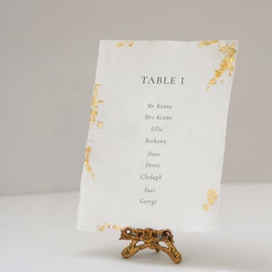May include: A white card with gold flecks on it, with the text "TABLE 1" and a list of names: Mr Keane, Mrs Keane, Ellie, Bethany, Dave, Denis, Clodagh, Suzi, George. The card is on a gold metal stand.