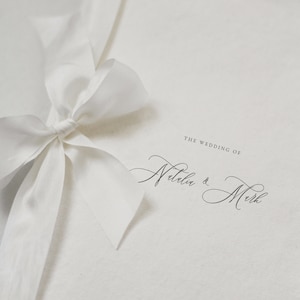 May include: White wedding invitation with a white satin ribbon tied in a bow. The invitation reads "The Wedding of Natalia & Mark" and "Saturday Nineteenth June".