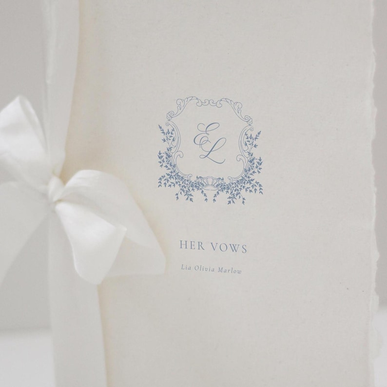 May include: A wedding vow booklet with a blue floral design and the text "HER VOWS". The booklet is tied with a white satin ribbon. The design includes the initials "E L" inside a decorative frame.
