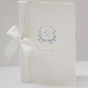 May include: A cream-colored wedding vow booklet tied with a white satin ribbon. The cover features a blue floral wreath with the initials "E L" and the text "HER VOWS" and the name "Lia Olivia Marlow".