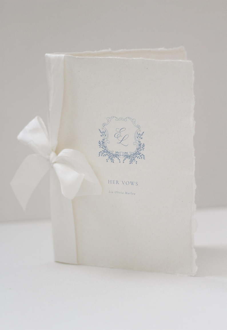 May include: A cream-colored wedding vow booklet tied with a white satin ribbon. The cover features a blue floral wreath with the initials "E L" and the text "HER VOWS" and the name "Lia Olivia Marlow".