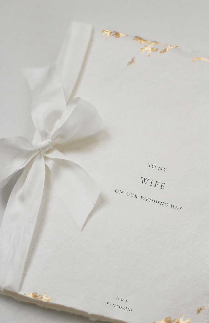 May include: A white card with gold accents and a white ribbon tied in a bow. The card says "To my Wife on our Wedding Day" and "A & J Santorini".