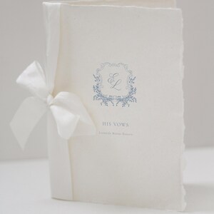 May include: A white wedding vow booklet tied with a white satin ribbon. The cover features a blue floral design with the initials "E L" and the words "HIS VOWS". The paper has a textured, deckled edge.