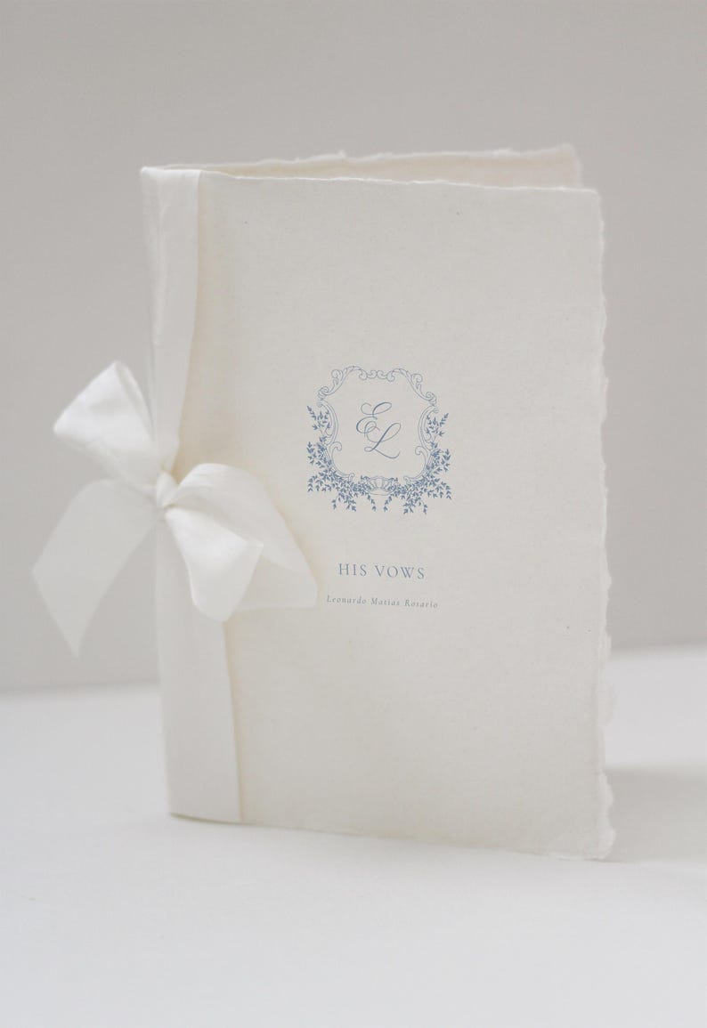May include: A white wedding vow booklet tied with a white satin ribbon. The cover features a blue floral design with the initials "E L" and the words "HIS VOWS". The paper has a textured, deckled edge.