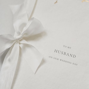 May include: A white card with gold accents and a white ribbon tied in a bow. The card says "To my Husband on our wedding day" and "A & J Santorini".