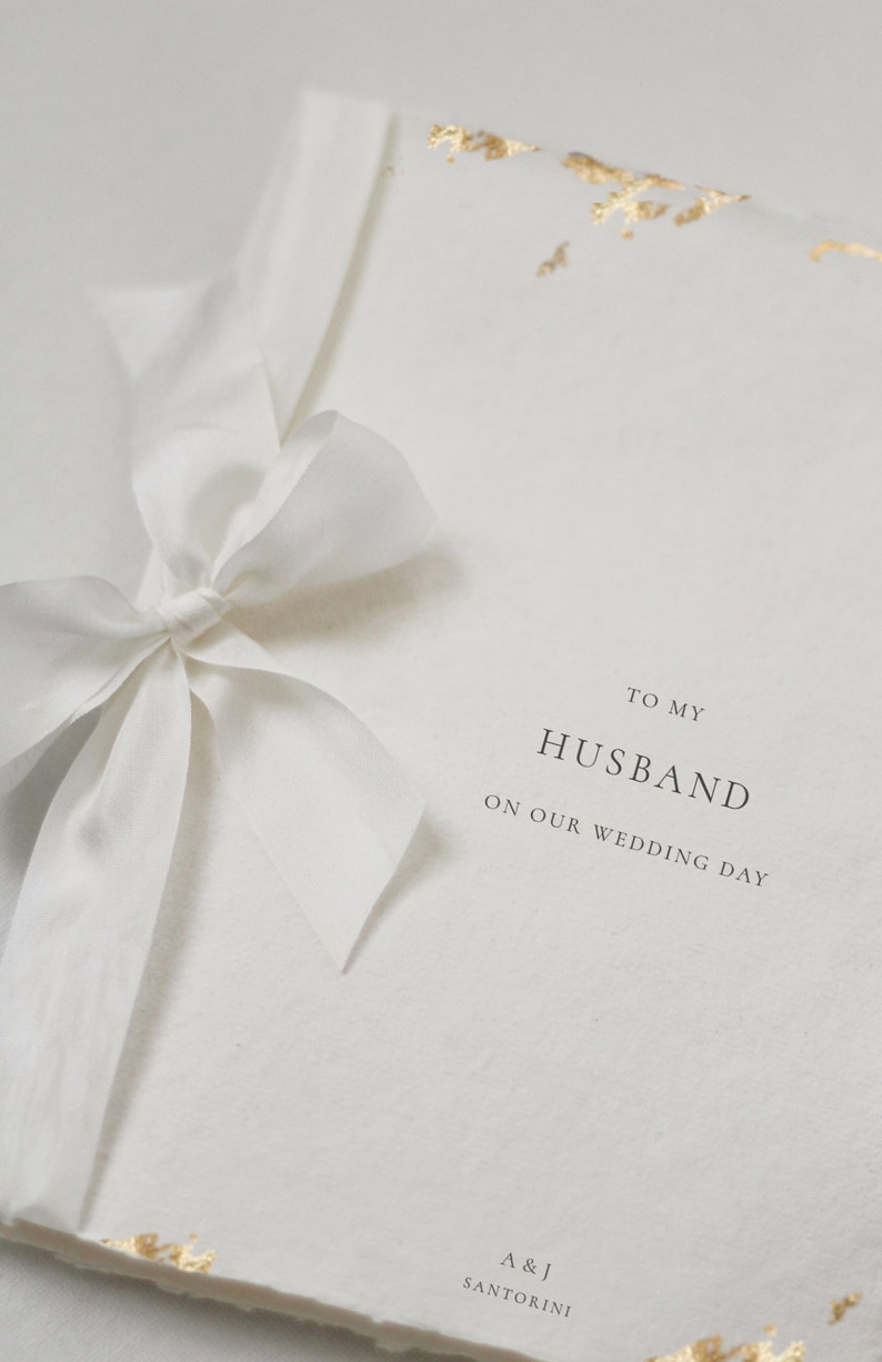 May include: A white card with gold accents and a white ribbon tied in a bow. The card says "To my Husband on our wedding day" and "A & J Santorini".