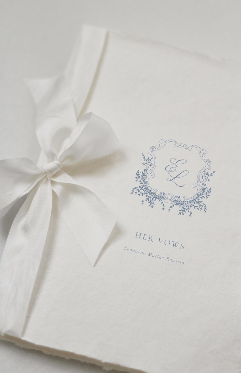 May include: A white wedding vow book with a light blue floral design and the text "HER VOWS". A white ribbon is tied in a bow on the left side of the book. The book is made of textured paper.