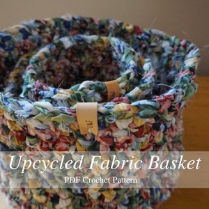 May include: A close-up of a crocheted fabric basket made from colourful scraps of fabric. The basket is made in a circular shape and has a layered design. The basket is labelled "Upcycled Fabric Basket PDF Crochet Pattern".