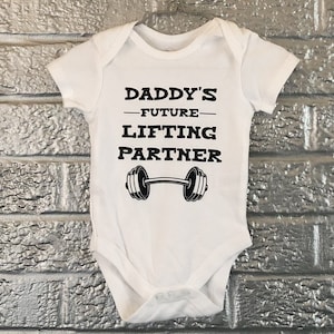 Peut inclure: Body bébé blanc avec l'inscription noire "DADDY'S - FUTURE - LIFTING PARTNER" et un haltère. Le body est suspendu à un cintre métallique devant un mur de briques argentées.