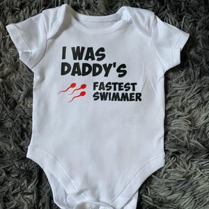 Puede incluir: Body de bebé blanco con mangas cortas. El texto "I WAS DADDY'S FASTEST SWIMMER" está impreso en negro, con tres gráficos de espermatozoides rojos. El body tiene un cierre de botón a presión en la parte inferior.