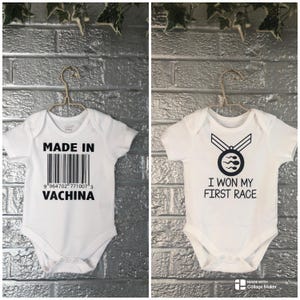 May include: Two white baby onesies hanging on a gold hanger. One onesie has the text "MADE IN VACHINA" with a barcode. The other onesie has a medal graphic and the text "I WON MY FIRST RACE".