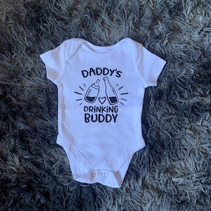 May include: White baby onesie with short sleeves. The onesie features the text "Daddy's Drinking Buddy" in black, along with illustrations of a baby bottle and a bottle with a heart. The onesie is made of soft material.