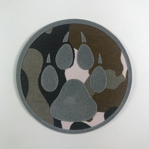 Könnte beinhalten: Ein runder, grauer Pfotenabdruck-Patch mit Tarnmuster-Hintergrund. Der Patch hat einen grauen Rand und ist aus Stoff.