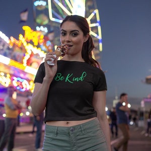 May include: A woman wearing a black crop top with the text "Be Kind" in green lettering, green shorts, and eating a chocolate ice cream cone. She is standing in front of a brightly lit Ferris wheel at an amusement park.