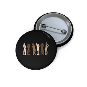 May include: A black button with a white background. The button features a sign language interpretation of the phrase "Be Kind" with hands of different skin tones.