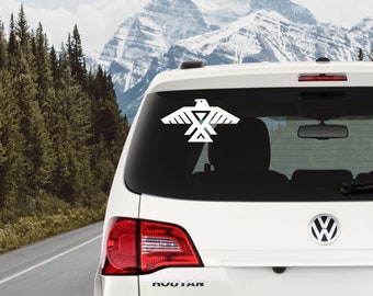 May include: White car with a white eagle decal on the back window. The decal is a stylized eagle with a geometric design. The car is parked on a road with mountains in the background.