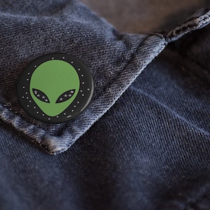May include: A black and green alien face button with white stars around the edge. The button is pinned to a blue denim jacket.