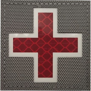 Könnte beinhalten: Ein grauer Stoff-Patch mit weißem Rand und einem roten reflektierenden Kreuz im Inneren. Das Kreuz hat ein Diamantenmuster.