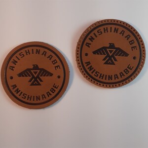 May include: Two brown leather patches with black stitching and the text "Anishinaabe" in a circle around a stylized bird design.