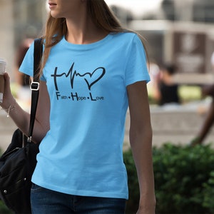 May include: A light blue t-shirt featuring a black graphic design of a cross, a heartbeat, and a heart. The text "Faith. Hope. Love." is printed below the graphic.