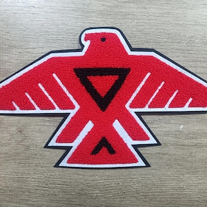 May include: A red and black embroidered patch in the shape of a stylized bird with outstretched wings. The patch features a black triangle and a white border. The patch is on a wooden surface.