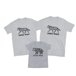 May include: Three grey t-shirts with wolf illustrations and text. The largest shirt says "Papa Wolf" and features a wolf walking to the right. The medium shirt says "Mama Wolf" and features a wolf walking to the right. The smallest shirt says "Little Wolf" and features a wolf cub walking to the right.