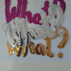 May include: Three iron-on patches featuring the word "What?" in different colours and styles. The patches are made of sequins and have a white backing. The first patch is pink, the second is silver, and the third is gold.