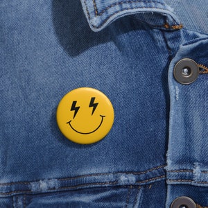 May include: Yellow smiley face button with black lightning bolts for eyes. The button is pinned to a blue denim jacket.