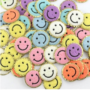 May include: A selection of colourful, round patches with embroidered smiley faces. The patches feature a variety of colours, including yellow, pink, blue, purple, orange, and white, each with a black smile and a gold trim. These patches are ideal for crafts.