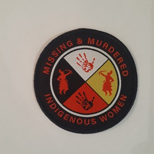 May include: A black and red circular patch with the text "Missing & Murdered Indigenous Women" around the edge. The center of the patch features a four-part design with a red handprint in each quadrant.