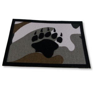 May include: A black bear paw print patch on a camouflage background. The patch is surrounded by a black border.