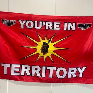 May include: A red flag with the words "YOU'RE IN MOHAWK TERRITORY" in white letters. A stylised sun with a Native American face in the centre is surrounded by yellow rays.
