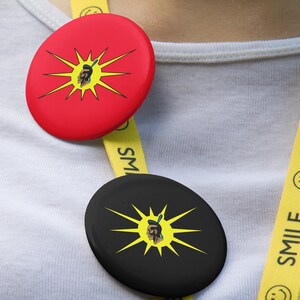 May include: Two round buttons with a yellow sunburst design and a black and white image of a person's face in the center. The buttons are attached to a yellow lanyard with the word "SMILE" printed on it.