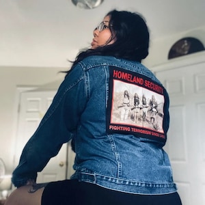 Könnte beinhalten: Eine blaue Jeansjacke mit einem Patch auf der Rückseite, auf dem "Homeland Security Fighting Terrorism Since 1962" steht und ein Schwarzweißbild von vier Personen mit Gewehren zu sehen ist.