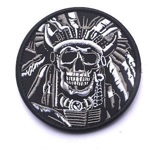 May include: Black and white embroidered patch featuring a skull wearing a headdress with feathers. The skull is intricately designed with a toothy grin.