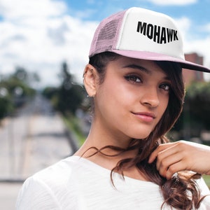 May include: A person wearing a white and pink trucker hat with black mesh and the word "MOHAWK" in black lettering. The hat has a black brim.
