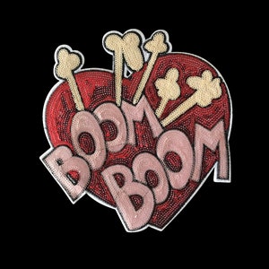 May include: A red heart-shaped patch with the word "BOOM" written in pink letters and surrounded by gold sequins. The patch has a glittery finish and is decorated with gold sequin stars.