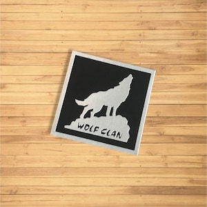 May include: A black square patch featuring a silver wolf silhouette howling at the moon. The text "WOLF CLAN" is printed below the wolf.