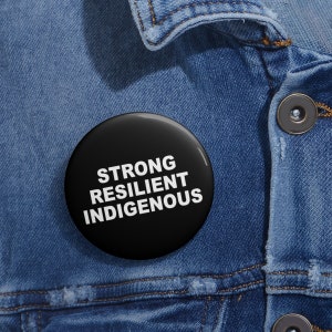 May include: Black button with white text that reads "STRONG RESILIENT INDIGENOUS" on a denim jacket.