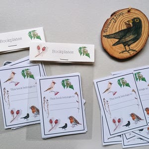 Peut inclure: Un ensemble d'ex-libris et un ornement en bois. Les ex-libris sont rectangulaires avec des illustrations à l'aquarelle d'oiseaux, de baies et de feuillage. L'ornement est une tranche de bois avec un oiseau peint et le texte "Raspberry Thief".