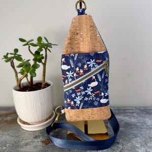 May include: A blue and white fabric crossbody bag with a cork bottom and a zipper closure. The bag features a pattern of white birds and flowers on a blue background. The bag has a long adjustable strap.