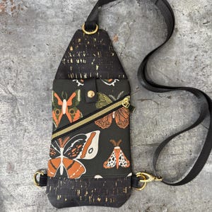 May include: A crossbody sling bag featuring a dark green fabric with a moth and butterfly print in orange, white, and black. The bag has black cork-like accents with gold flecks, a diagonal gold-tone zipper pocket, and an adjustable black strap.