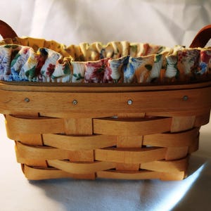 May include: A small, oval-shaped, wooden basket with two handles. The basket is lined with a floral fabric with a white background and colorful flowers.