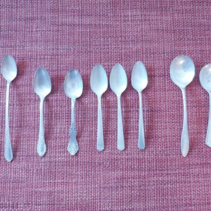 May include: A set of nine silver spoons, arranged in a row on a pink textured surface. The spoons are all different sizes and shapes, with some having ornate handles.