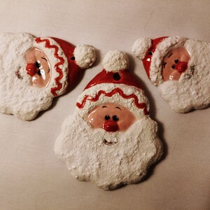 May include: Three ceramic Santa Claus ornaments with white fuzzy beards and red hats. The ornaments have painted faces with black eyes, red noses, and pink cheeks.