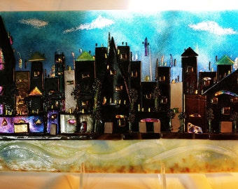 May include: A glass art piece depicting a cityscape with a blue sky and white clouds. The buildings are black and have gold accents. The cityscape is set against a background of a wavy, light blue and green water.