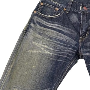 Puede incluir: Primer plano de vaqueros de mezclilla azul desgastados. Los vaqueros tienen un lavado descolorido con desgaste visible, incluyendo parches claros y un &aacute;rea desgastada cerca de la rodilla. Incluye remaches met&aacute;licos y un dise&ntilde;o cl&aacute;sico de cinco bolsillos.